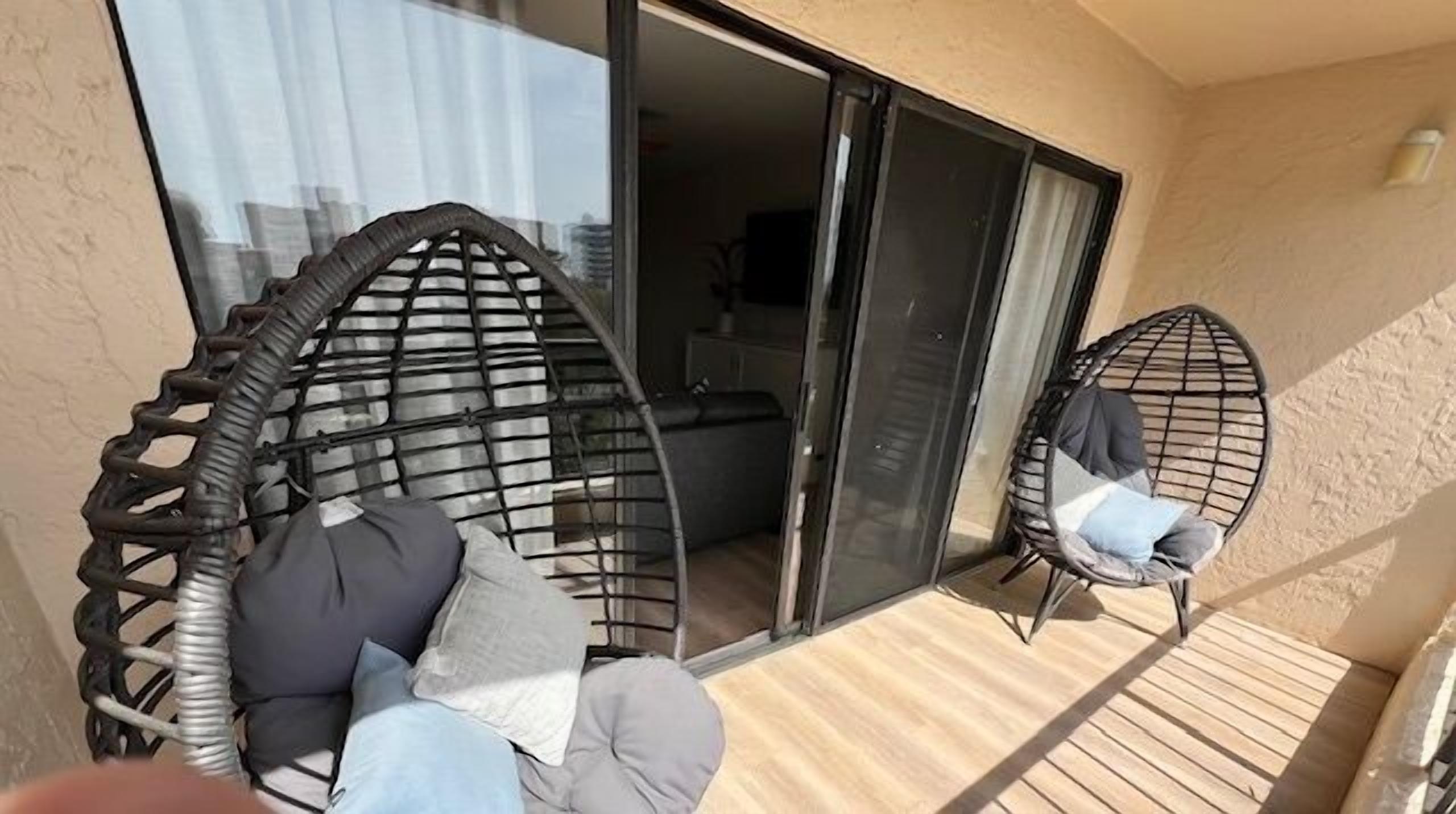 Een balkon met twee rieten stoelen, waarvan er één kussens heeft, en een schuifdeur naar een binnenruimte.