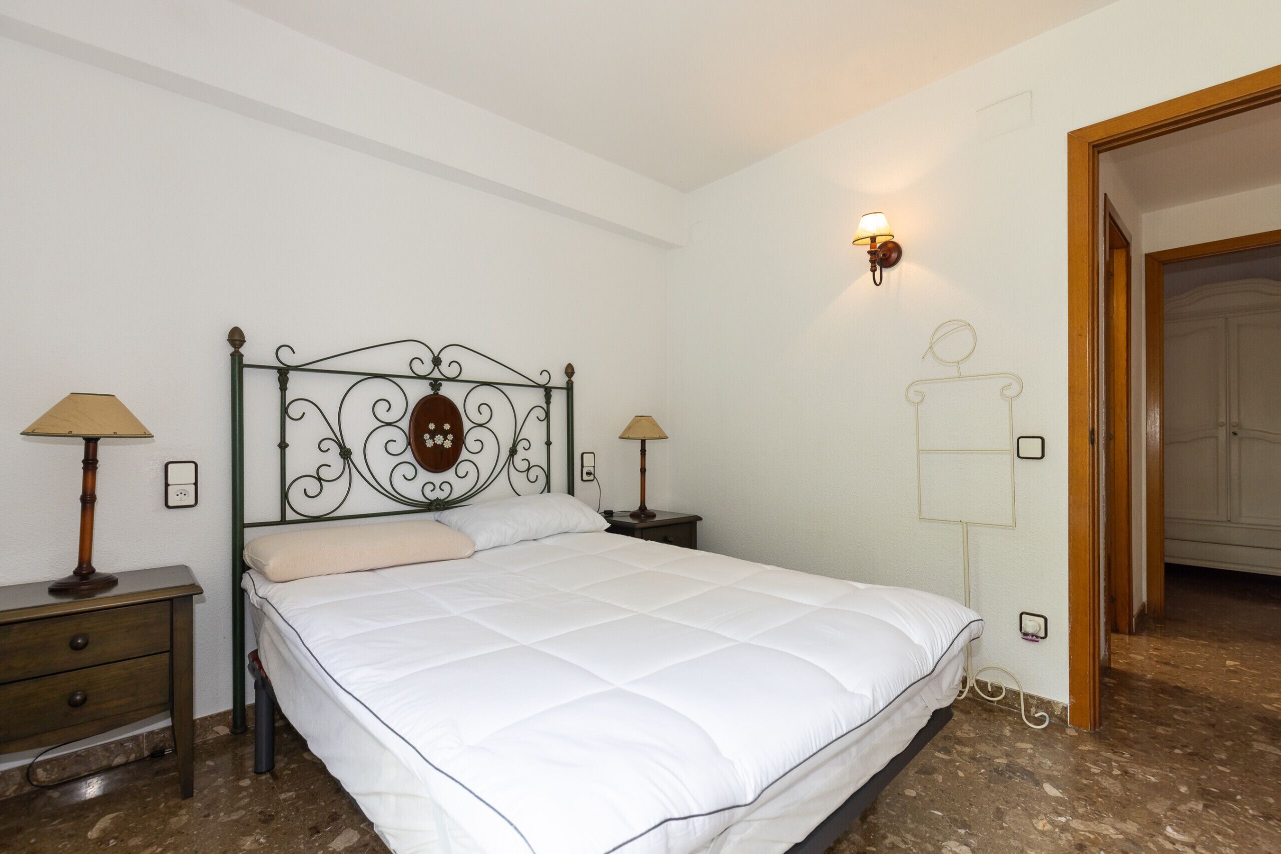 A bedroom with a bed, two bedside tables, a wall-mounted lamp, and a door leading to another room.