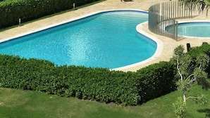 A large, curved swimming pool surrounded by a sandy area and dense green hedges.