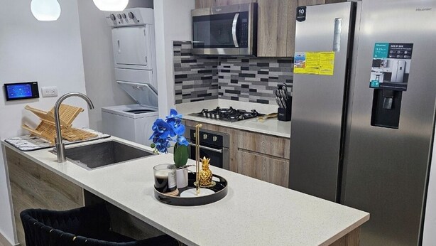 A modern kitchen with a central island, black bar stools, stainless steel appliances, and pendant lighting.