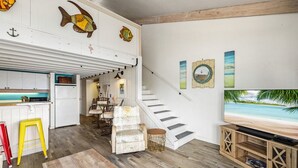 Un vaste espace de vie doté d’un plancher en bois, d’un escalier, d’une cuisine avec des meubles blancs et d’un téléviseur diffusant une scène de plage.