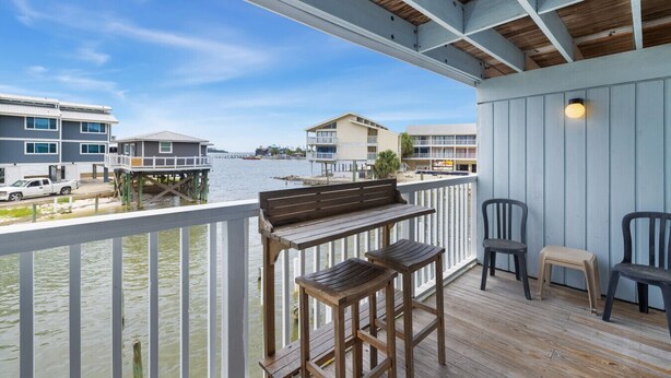 A wooden deck with a bench and chairs overlooking a waterfront with buildings in the background.