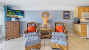 A room with two wicker chairs with orange cushions, a small table with a lamp, a wicker cabinet, a television, a framed picture, and a kitchenette.