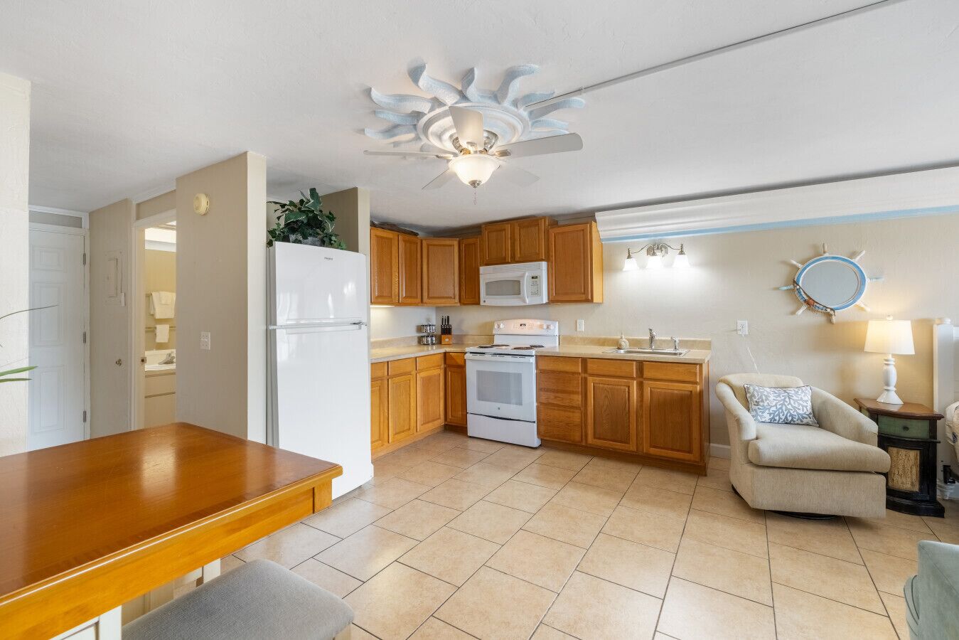 A compact kitchen with wooden cabinets, a white refrigerator, a stove, and a microwave. There's a dining table with a bench, a chair, and a small side table with a lamp.