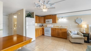 A compact kitchen with wooden cabinets, a white refrigerator, a stove, and a microwave. There's a dining table with a bench, a chair, and a small side table with a lamp. - Balcony 🌴 King Bed + Sofa Bed 🌴 Water Views (Cedar Key)