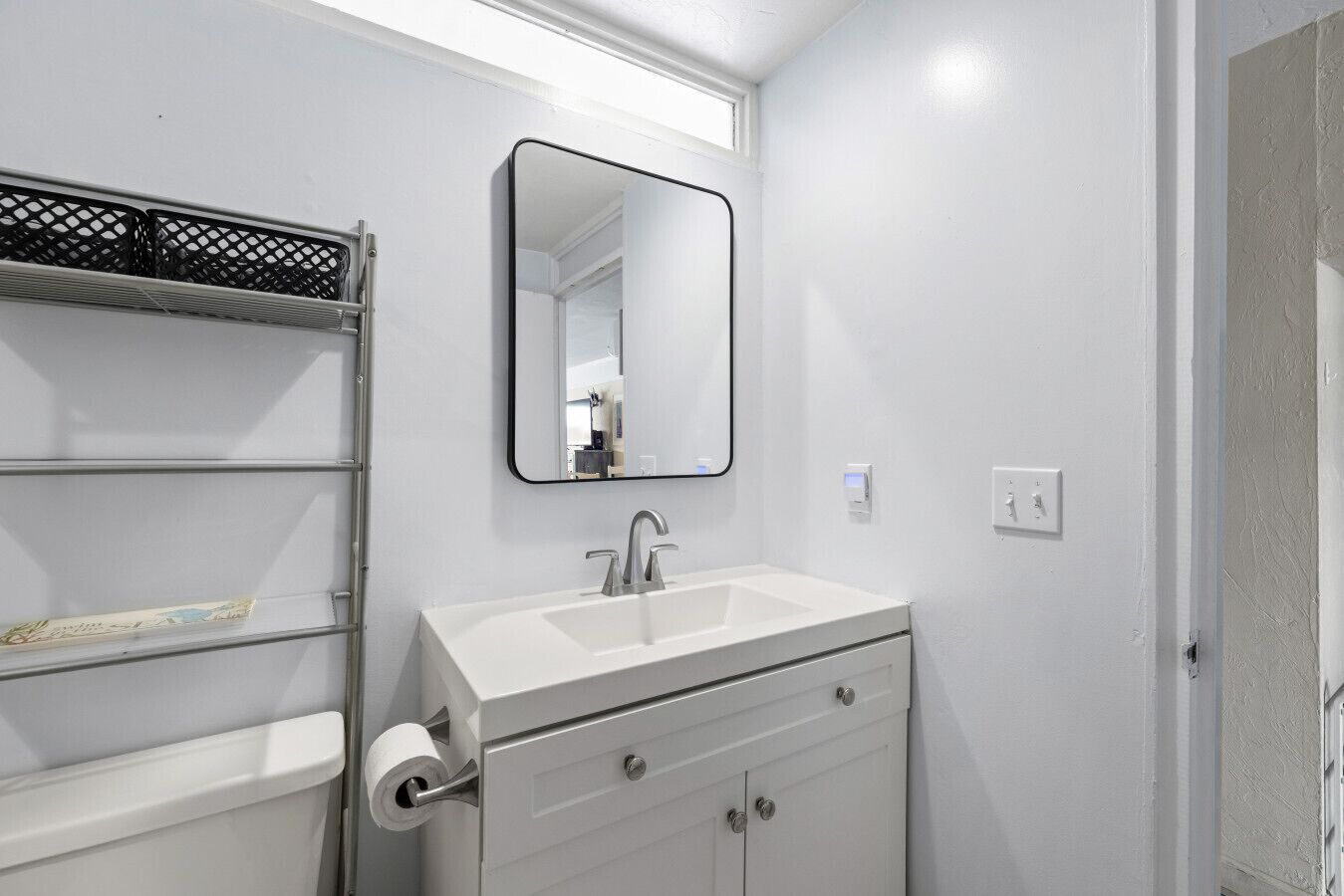 A bathroom with a white sink, a mirror, a toilet, and a shelf with a roll of toilet paper.