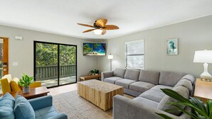 A living room with a grey sofa, blue armchair, wooden coffee table, and a ceiling fan. There is a sliding glass door leading to a balcony with a view of trees. - A Cozy Canopy Getaway in Cedar Key (Cedar Key)