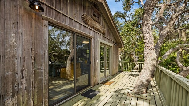 A wooden deck with a glass door leading to a covered area, surrounded by trees and a wooden fence. - A Cozy Canopy Getaway in Cedar Key (Cedar Key)