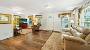 A spacious living room with a leather sofa, a dining area with a table and chairs, and a kitchen area with a refrigerator and a window.