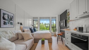 A modern apartment with a large bed, a sofa, a kitchen with a TV, and a view of greenery.