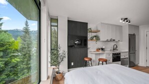 A modern bedroom with a large bed, a small kitchen, and a view of a forest.