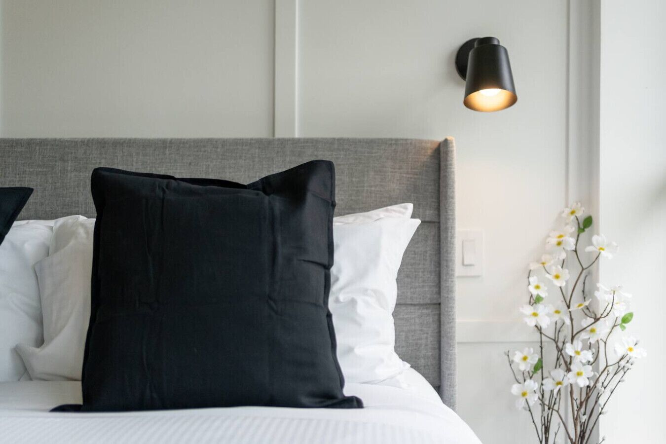 Una cama bien hecha con almohadas blancas y negras, un cabecero gris y una lámpara fijada a la pared.