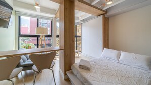 A modern bedroom with a large bed, a desk, and a chair, featuring a view of the city through large windows.