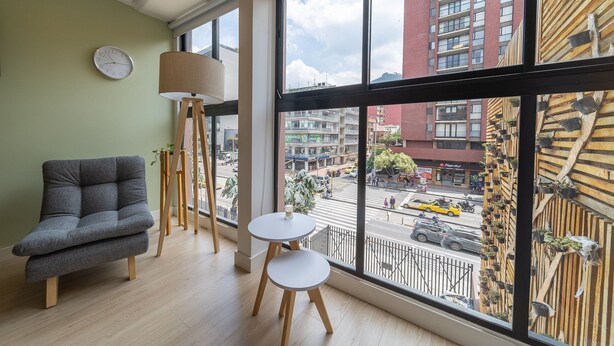 A modern room with a sofa, two small tables, a floor lamp, and a large window offering a view of a busy street and buildings.