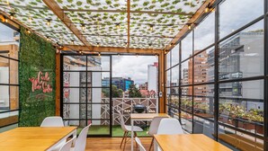 A modern cafe with wooden tables and white chairs, large windows, and a green wall with a neon sign.