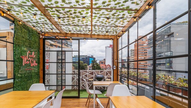 A modern cafe with wooden tables and white chairs, large windows, and a green wall with a neon sign. - |Ahead| Spacious 3BR @Chapinero with Co-working (Bogotá)