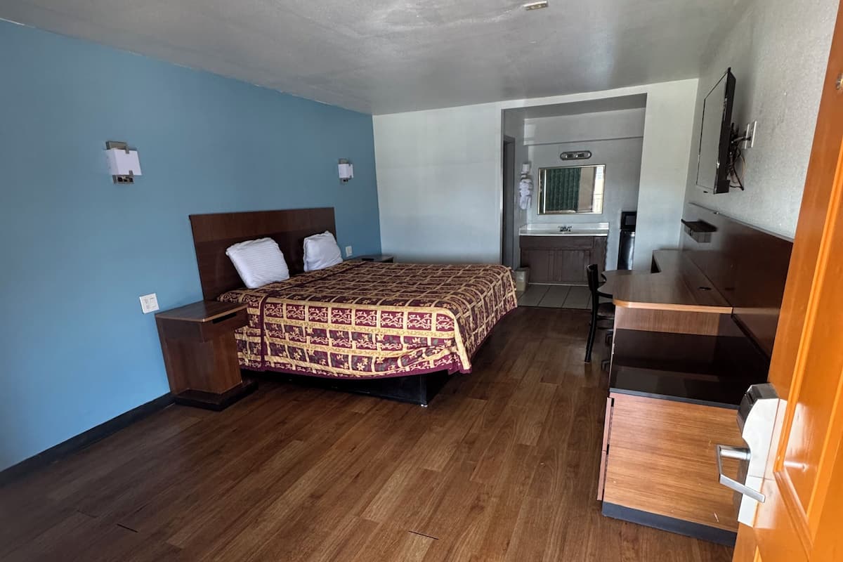 Habitación de hotel con piso de madera, una cama con colcha estampada, un escritorio, una silla y una pequeña zona de kitchenette.