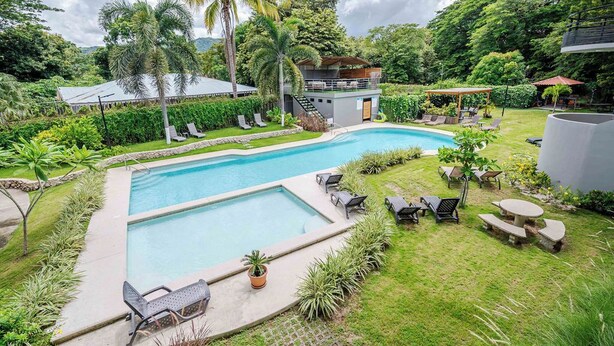 Une piscine dotée de chaises longues et d’un espace de repos couvert, le tout entouré d’une végétation luxuriante et d’un petit bâtiment.
