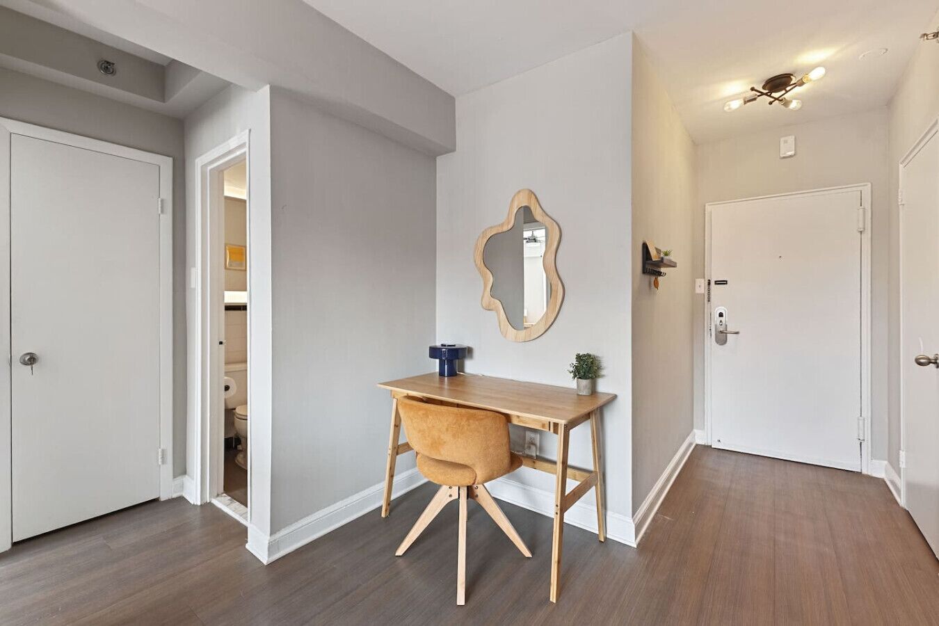 A hallway with a wooden desk and chair, a mirror on the wall, and a door leading to a bathroom.