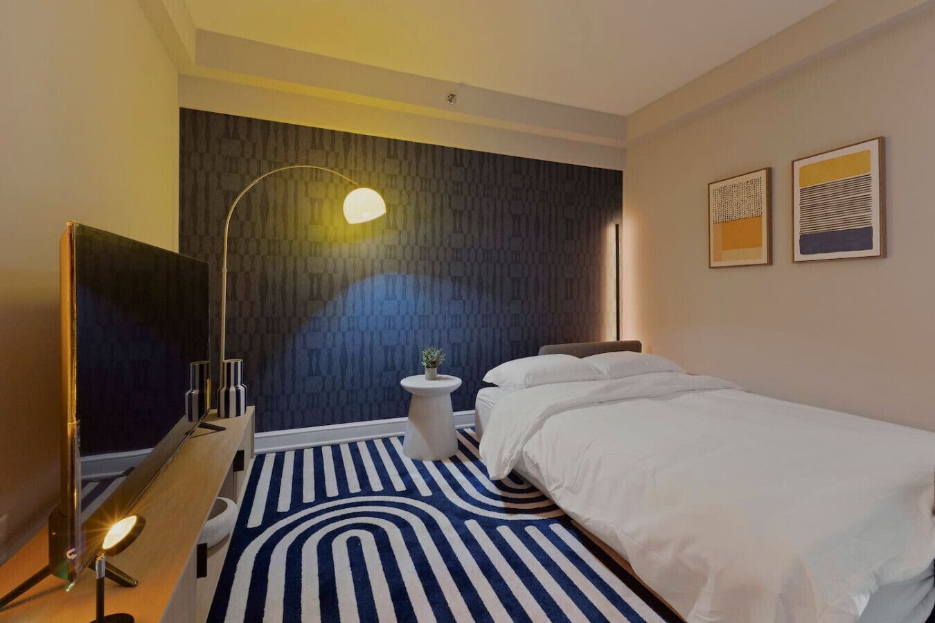 A modern hotel room with a large bed, a desk, a lamp, and a small table with a plant.