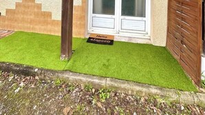 Ein Haus mit einer Holzveranda, einer Doppeltür mit Glasfenster und einem kleinen Garten mit Kunstgrashalm.
