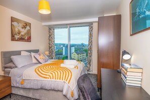 Une chambre d’hôtel équipée d’un lit, d’un bureau, d’une chaise, d’une lampe et offrant une vue sur la ville grâce à de grandes fenêtres.