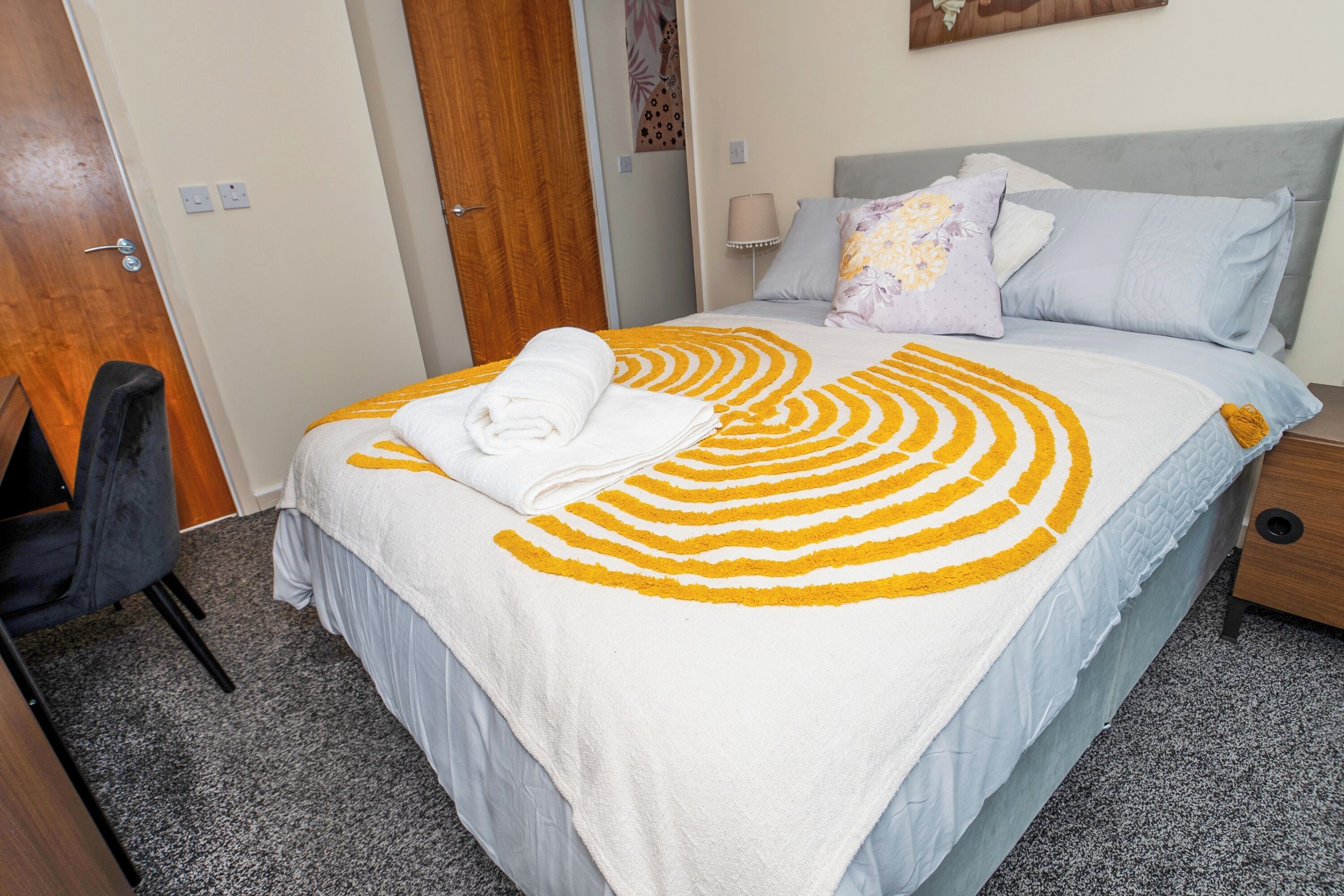 A neatly made bed with a white and yellow striped bedspread, a grey headboard, and a bedside table with a lamp.