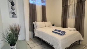 Un dormitorio con cama, ventilador de techo, ventana con cortinas, una obra de arte en la pared y una mesita con planta.