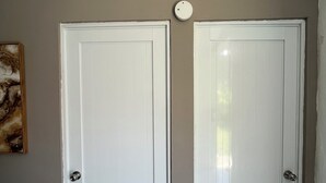 Dos puertas blancas con un detector de humo encima, integradas en una habitación con una pared lisa y suelo de baldosas.