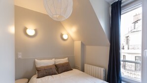 A bedroom with a bed, two wall-mounted lights, a radiator, and a window with curtains. - The (Courdemanche)