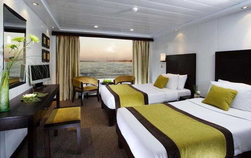 A hotel room with two beds, a desk, and a chair. There is a view of the sea through the window.