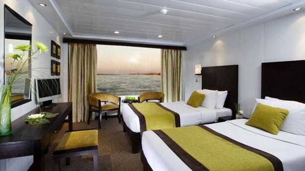A hotel room with two beds, a desk, and a chair. There is a view of the sea through the window. - Movenpick MS Royal Lily Nile Cruise (Luxor)