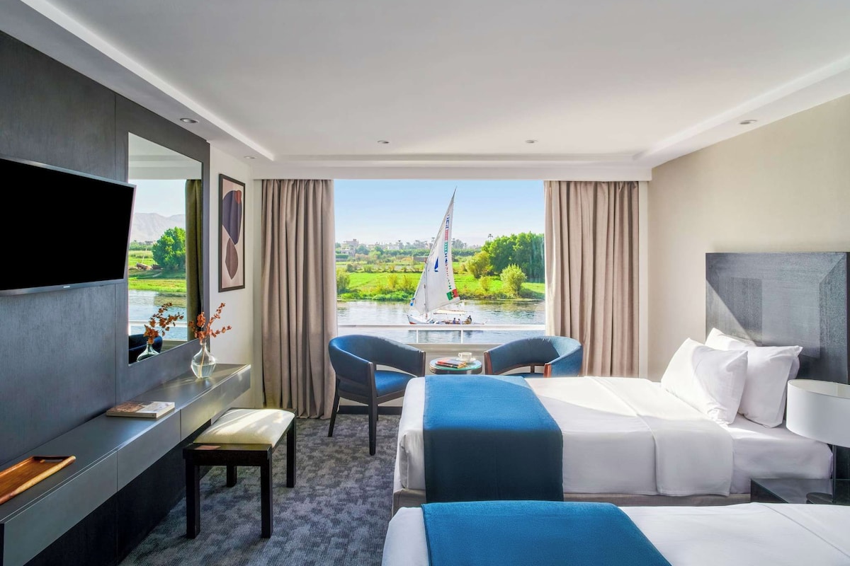 A hotel room with a bed, a desk, a chair, a TV, and a view of a river with a sailboat.