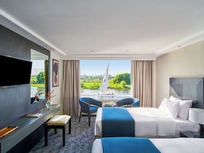 A hotel room with a bed, a desk, a chair, a TV, and a view of a river with a sailboat. - Movenpick MS Royal Lily Nile Cruise (Luxor)