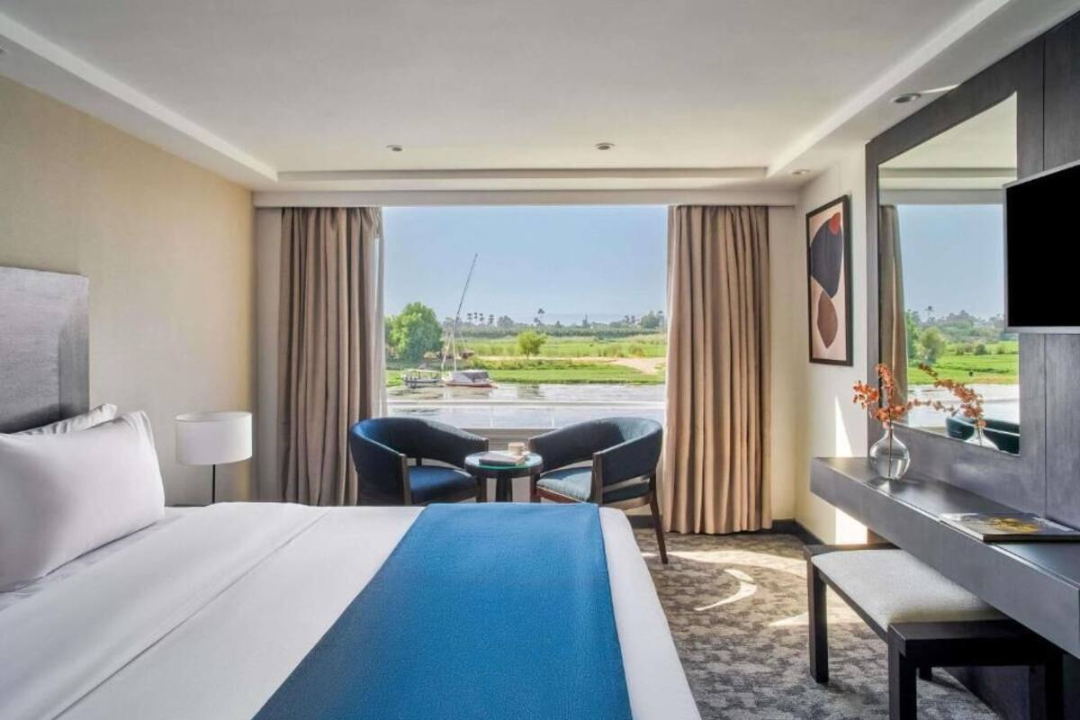 A hotel room with a large bed, a desk, a chair, and a view of a green landscape.
