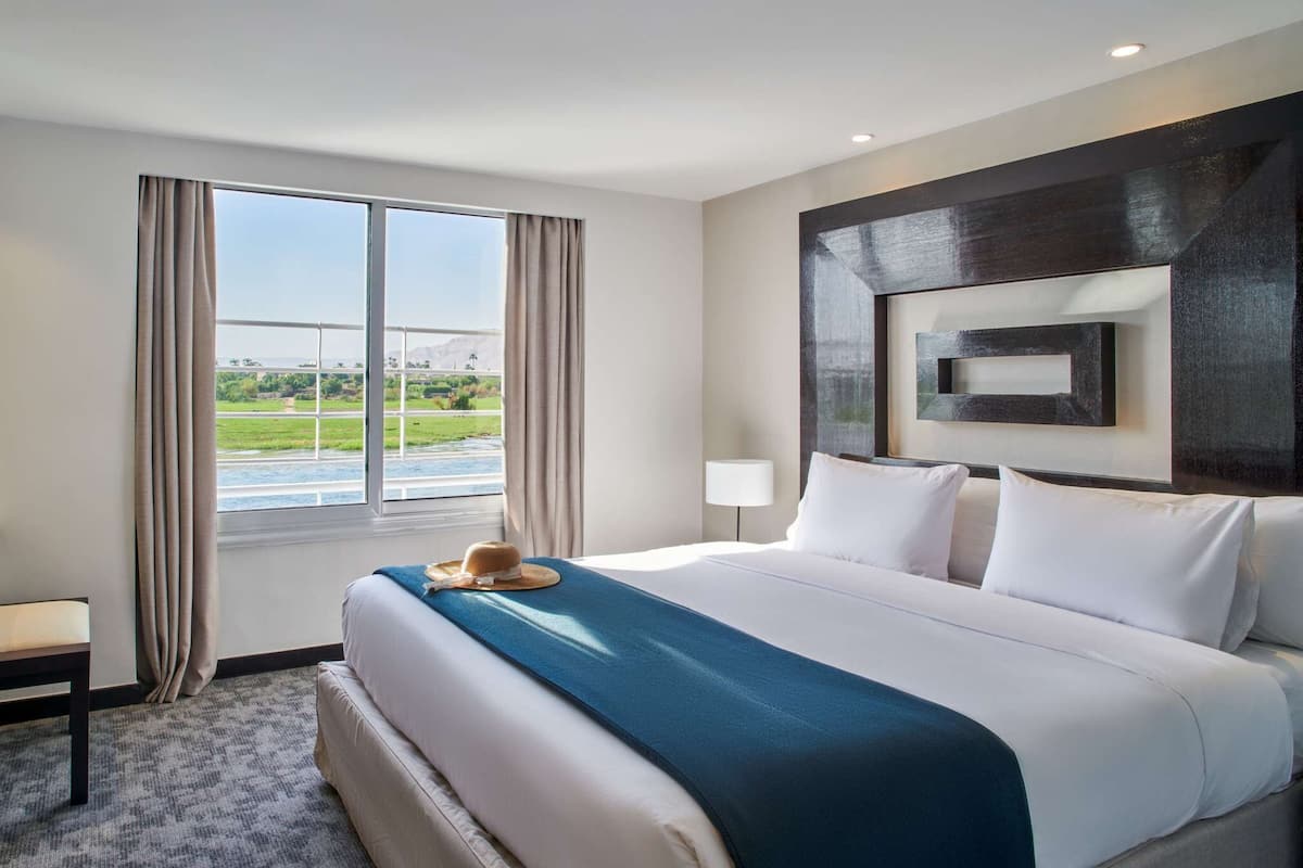 A hotel room with a large bed, a view of a river and greenery, and a modern wall design.