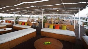 Un espacio exterior cubierto con sofás acolchados y mesas redondas, con vistas a una masa de agua.