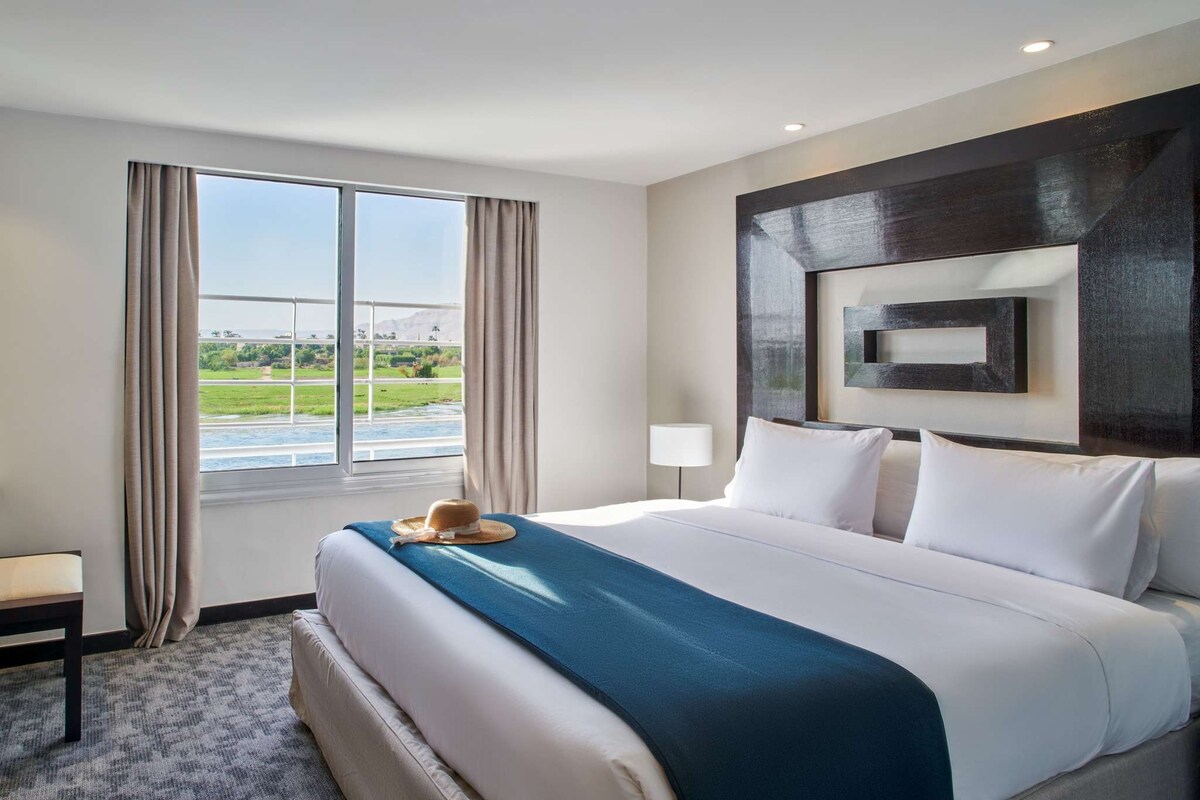 A hotel room with a large bed, a view of a river and greenery, and a modern wall design.