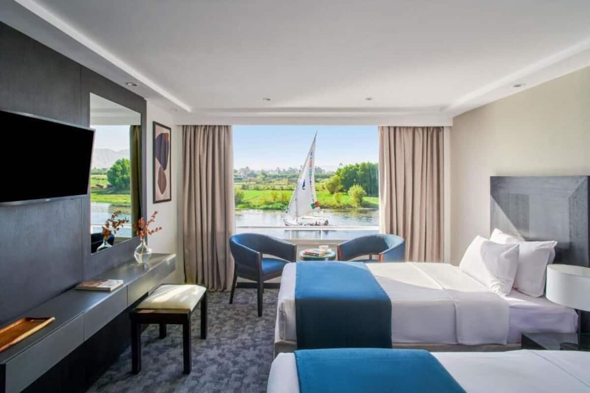 A hotel room with a bed, a desk, a chair, a TV, and a view of a river and greenery.