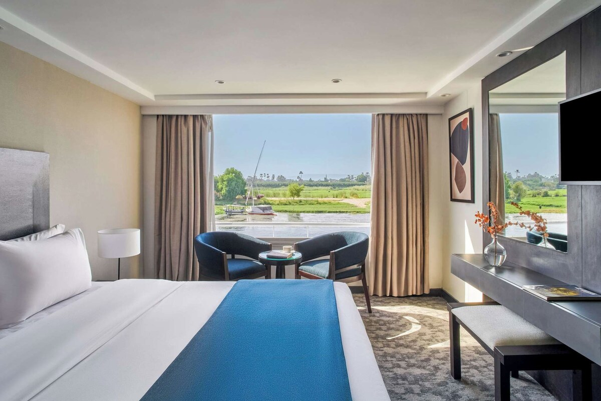 A hotel room with a bed, a desk, a chair, and a view of a river and boats.