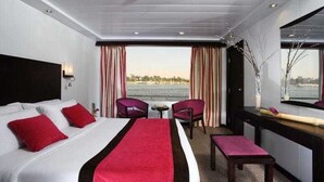 A hotel room with a bed, a desk, two chairs, a small table, and a view of the water. - Movenpick MS Royal Lily Nile Cruise (Luxor)