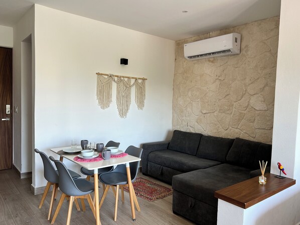 A modern living room with a dining area, stone wall, macramé wall hanging, and a small table with chairs. - Bohemian Apartment, Rooftop Pool with Ocean View! (Playa del Carmen)