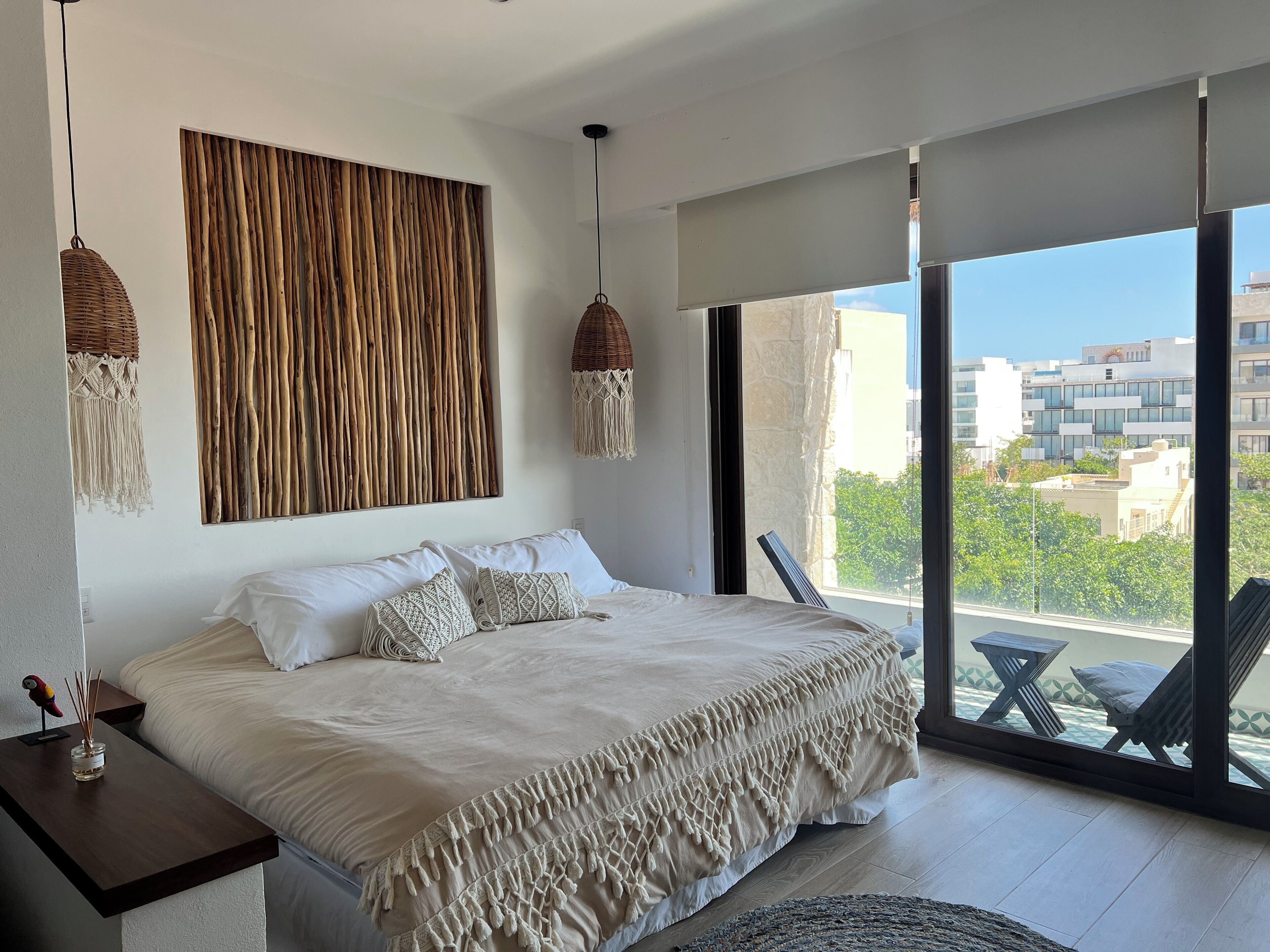 A modern bedroom with a large bed, macrame wall hanging, and a view of the city through large windows.