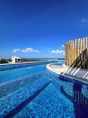 A swimming pool with blue mosaic tiles, a wooden fence, and a view of the ocean and buildings in the background. - Bohemian Apartment, Rooftop Pool with Ocean View! (Playa del Carmen)