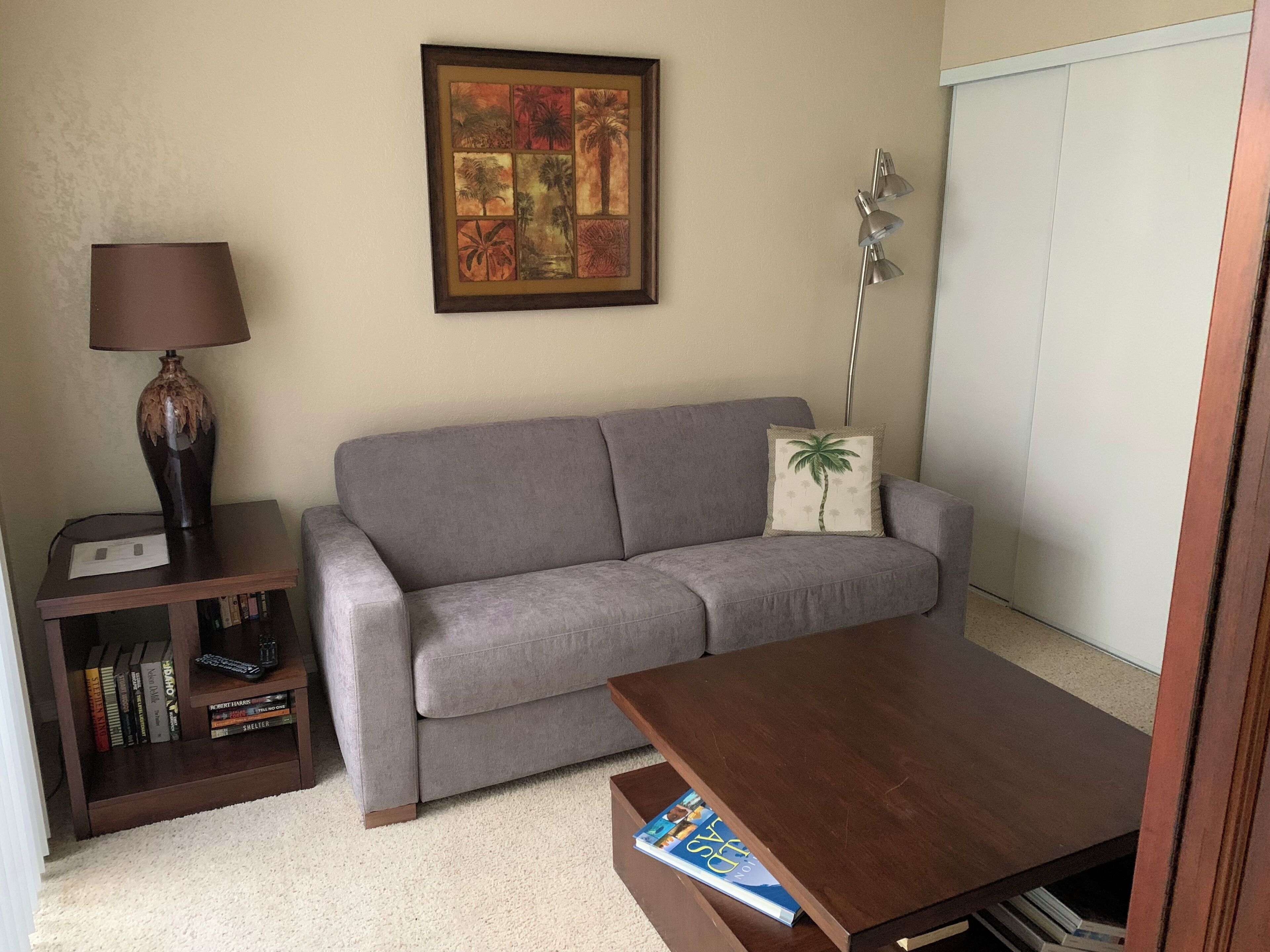 A living room with a grey sofa, a small wooden table, a bookshelf, a lamp, and a framed artwork on the wall.