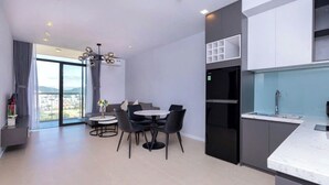 A modern kitchen with a marble countertop and a built-in oven. There is a dining area with a round table and chairs. The room has large windows with a view of the city. - Premium CSJ Apartment - Nice View and Walk To Vung Tau Beach (Vũng Tàu)
