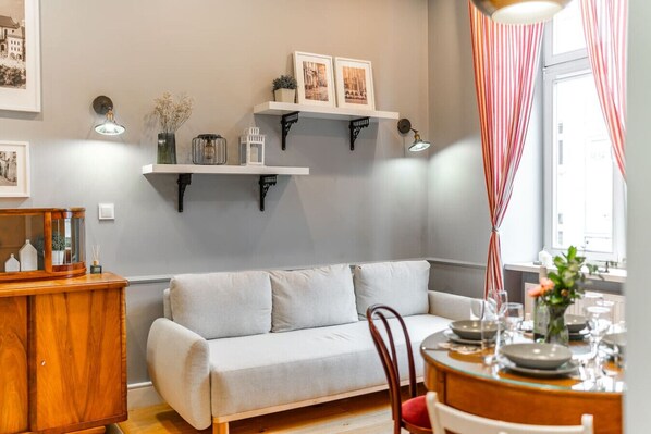 A living room with a sofa, a wooden side table, a dining table set with plates and glasses, and a window with curtains.