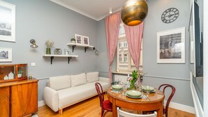 A dining area with a round wooden table set for four, a white sofa, and a red chair. There are two wall-mounted shelves with decorative items and a clock.