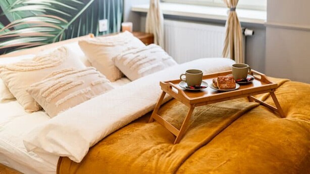 A bed with a wooden tray holding a cup and a pastry, a window with sheer curtains, and a wall with a large leaf pattern.