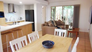 A modern kitchen with a central island, a dining area with a wooden table and white chairs, and a living area with a sofa and a TV. - Wallaby Hideaway (Trinity Beach)